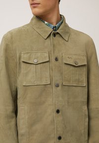 Veste en daim vert olive avec col, deux poches à rabat à l'avant, fermetures à pression et texture lisse. Détails de couture notables visibles.