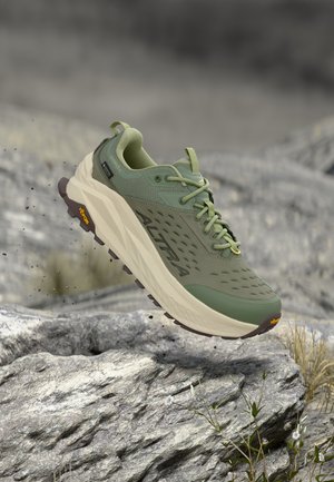 Chaussure de trail de course verte et beige en l'air au-dessus d'un terrain rocheux, mettant en avant une semelle épaisse et une adhérence texturée pour une utilisation en extérieur.