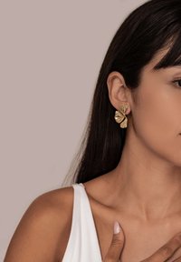 Mujer con cabello largo y oscuro, vistiendo una blusa sin mangas blanca y pendientes en forma de concha dorada, con la mano descansando cerca de la clavícula sobre un fondo claro.