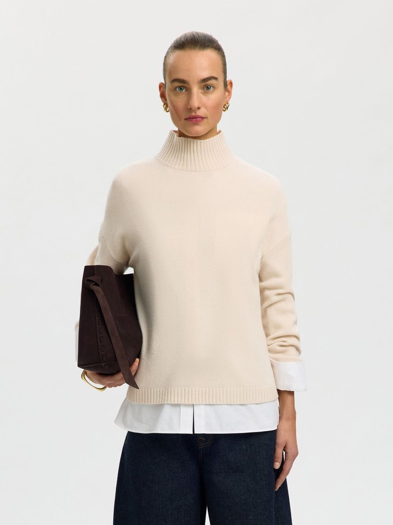 Femme portant un pull à col roulé beige clair, une chemise blanche en dessous, un pantalon foncé, tenant un sac à main marron foncé, debout devant un fond uni.