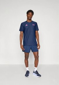 Souprava sportovního oblečení v námořnické modři, obsahující tričko s krátkým rukávem a odpovídající šortky. Součástí jsou růžové logo akcenty a látka odvádějící vlhkost.