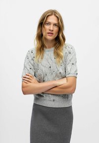 Pull en maille gris clair à manches courtes, avec un motif en torsades et des perles décoratives. Porté avec une jupe grise ajustée.