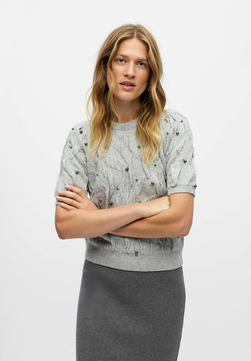 Pull en maille gris clair à manches courtes, avec un motif en torsades et des perles décoratives. Porté avec une jupe grise ajustée.