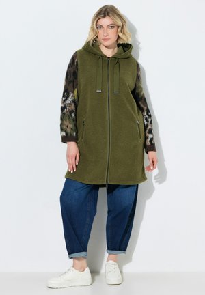 Kvinde iført blå jeans, hvide sneakers, en grøn ærmeløs hættetrøje med lynlås og en leopardprintet langærmet top.