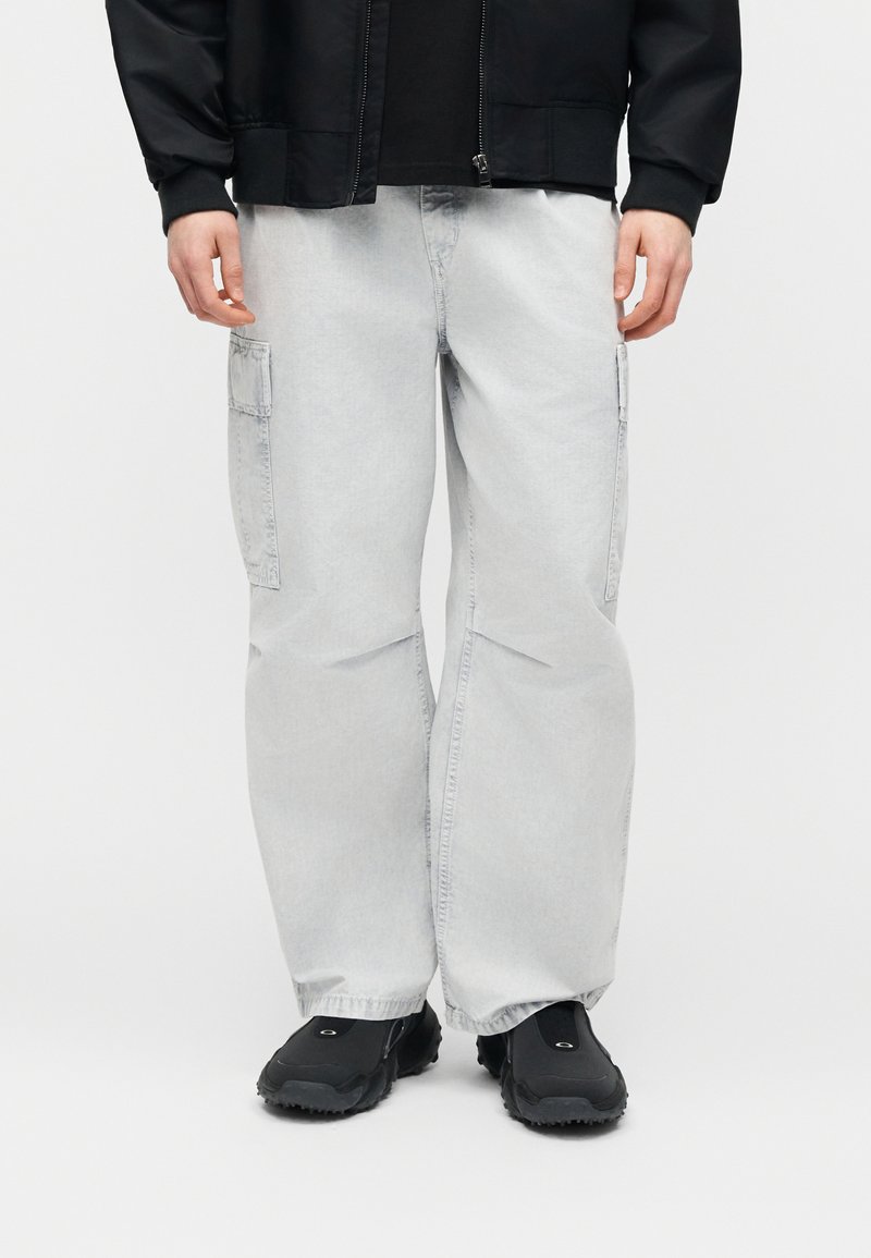 Person trägt hellgraue, weit geschnittene Cargohosen mit seitlichen Taschen, schwarze Schuhe und eine schwarze Jacke mit sichtbaren Bündchen.