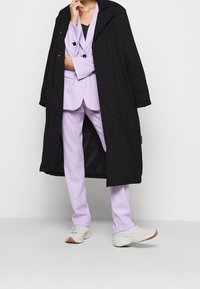 Schwarzer langer Mantel über einem helllila Anzug, der aus einer Jacke und Hose besteht, kombiniert mit weißen Sneakers, im entspannten Schnitt und mit glatter Textur.