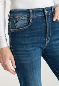 Jean bleu taille haute en denim. Les caractéristiques incluent des poches avant, un léger délavage et des accents de couture en marron contrasté.