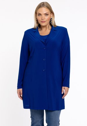 Frau in einem langen blauen Blazer mit Knöpfen über einem passenden Oberteil und blauen Jeans, mit hängenden herzförmigen Ohrringen.