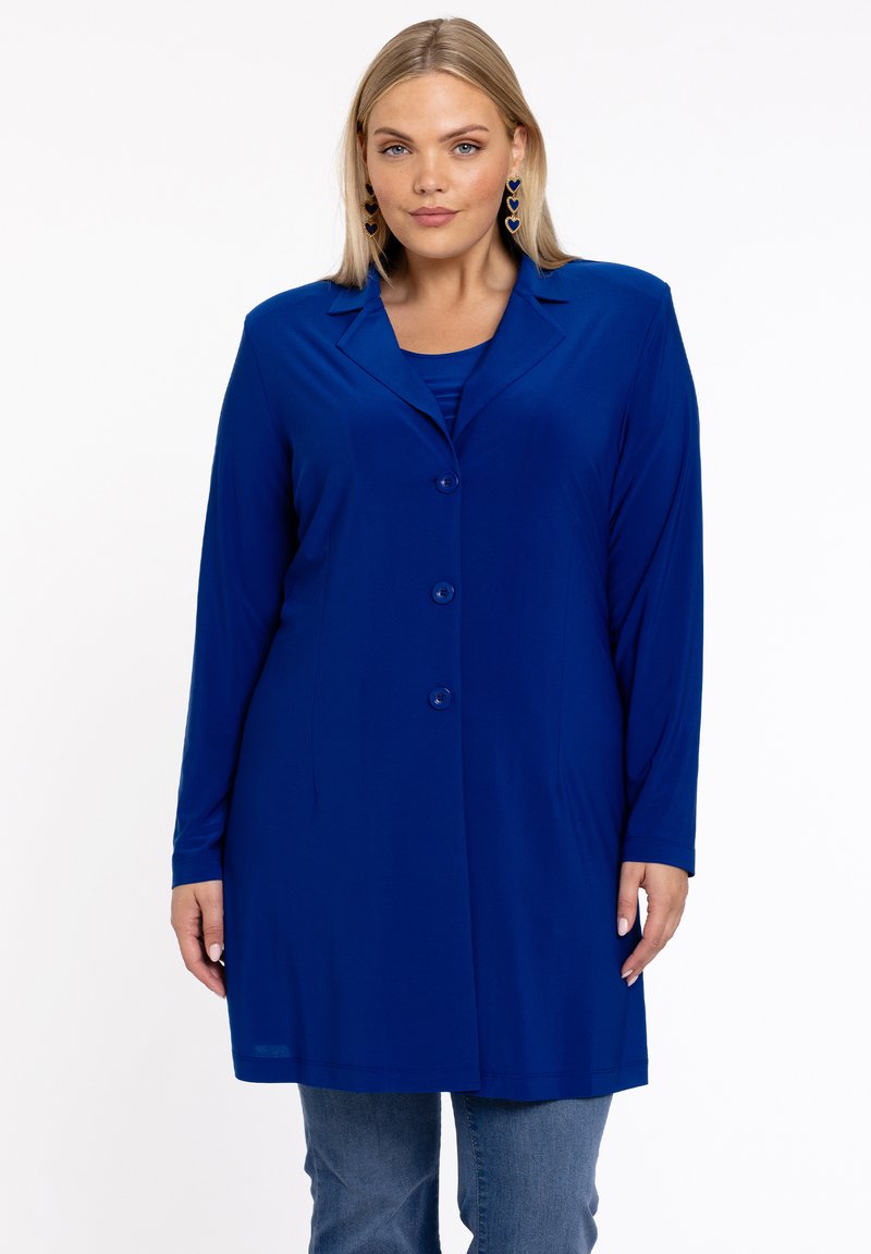Frau in einem langen blauen Blazer mit Knöpfen über einem passenden Oberteil und blauen Jeans, mit hängenden herzförmigen Ohrringen.