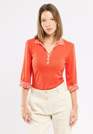 Femme aux cheveux mi-longs portant un haut orange avec un col et des poignets à fleurs, assorti à un pantalon crème taille haute.