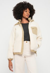 Chaqueta blanca forrada de forro polar con detalles en color cuero, cremallera frontal, dobladillo ajustable y un bolsillo en el pecho. Se lleva puesta sobre una camiseta negra y pantalones crema.