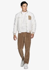 Hvid uld varsity jakke med brune detaljer, parret med brune fløjl bukser. Outfitten indeholder en hvid sweatshirt og hvide sneakers, mens der holdes en baseball.