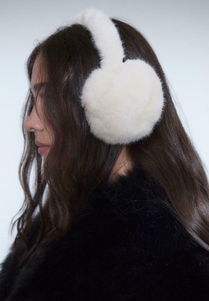 Faux fur earmuffs in cream color with a soft, round design and a curved band for secure wear, set against dark hair and clothing.