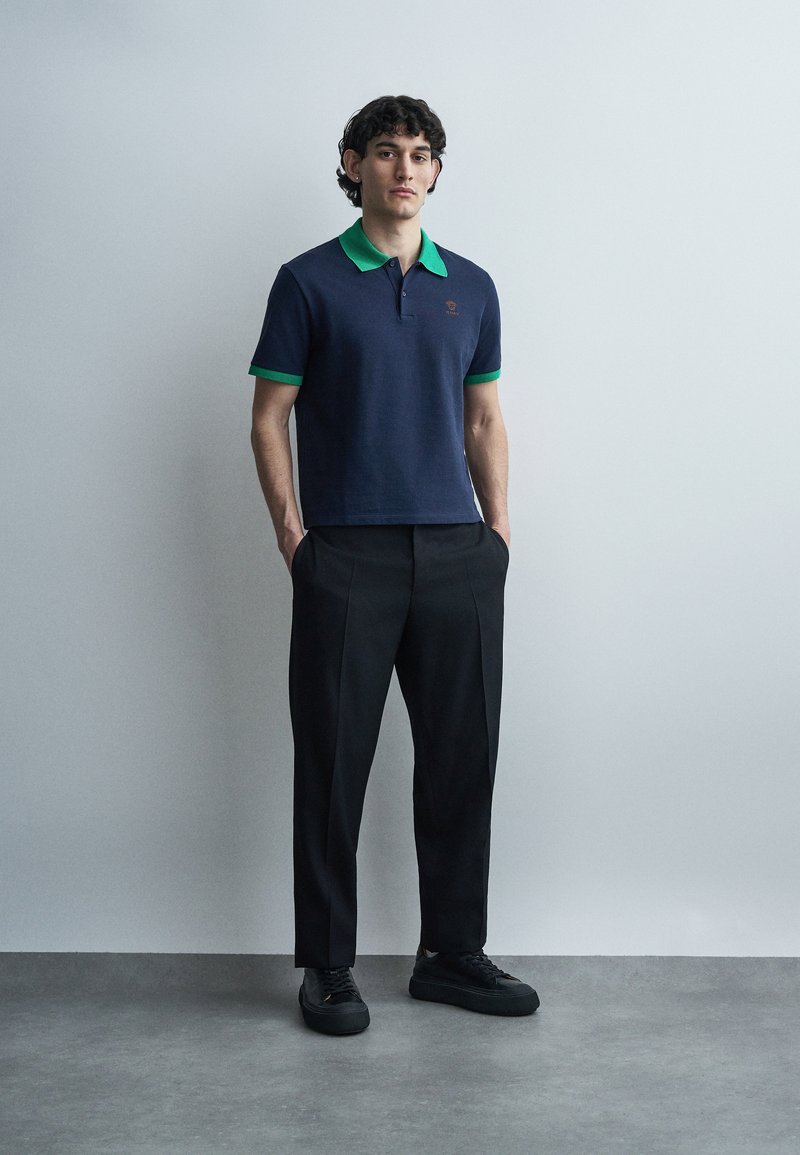 Jonge man staand met handen in de zakken, gekleed in een navy poloshirt met groene kraag en manchetten, zwarte broek en zwarte sneakers.