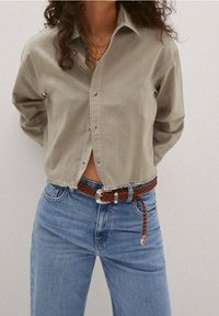 Camicia button-up grigia chiara con orlo leggermente accorciato, indossata con jeans in denim blu e una cintura intrecciata marrone con fibbia in argento.