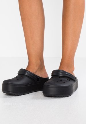 Jambes portant des sabots à plateforme noirs avec des trous de ventilation et des sangles de talon réglables sur un sol clair avec un fond blanc.