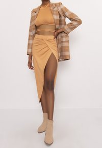 Top bodycon beige y falda envuelta con una gran abertura, combinados con una chaqueta de cuadros y botines beige, creando un look coordinado.