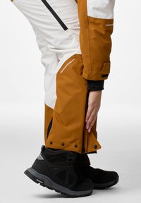 Waterproof ski pants in brown and white, featuring a zippered leg opening, black accents, and a durable, textured fabric. Black hiking boots visible.