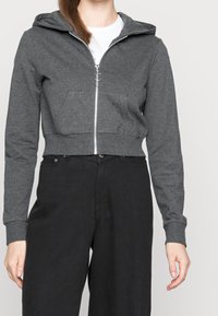 Sudadera gris recortada con cremallera y bolsillos frontales, hecha de tela de sudadera. Presenta una capucha y puños acanalados. Combinada con pantalones negros.