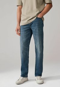 Jeans din denim de culoare albastră deschisă, cu croială dreaptă, ușor decolorate și design cu cinci buzunare, asortate cu o cămașă bej deschis și adidași albi.