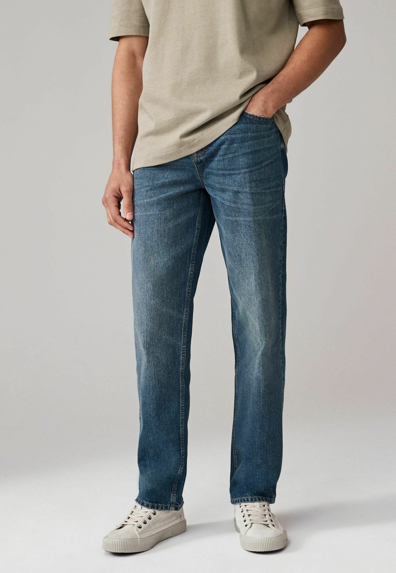 Jeans din denim de culoare albastră deschisă, cu croială dreaptă, ușor decolorate și design cu cinci buzunare, asortate cu o cămașă bej deschis și adidași albi.