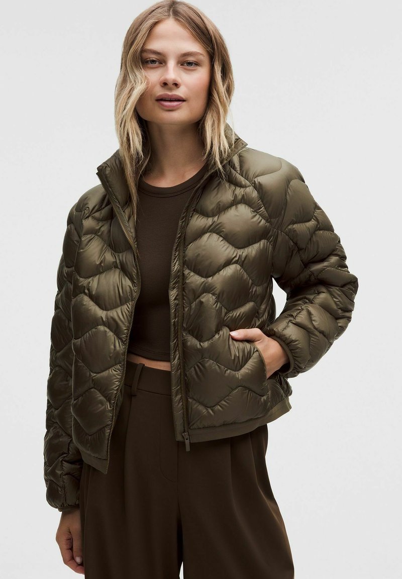 Młoda kobieta w oliwkowozielonej pikowanej kurtce puchowej nosi ciemnoskórą crop top i brązowe spodnie z wysokim stanem, stoi na tle jednolitego tła.