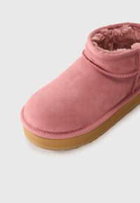 Bottine en daim rose avec doublure en fourrure et semelle en caoutchouc marron sur un fond blanc.