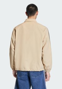 Beige lichtgewicht jack met een ontspannen pasvorm, elastische manchetten en een zwart zijaccent. Glad textuur en minimalistisch design. Achteraanzicht zichtbaar.