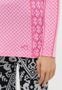 Langermet topp i rosa med et strukturert geometrisk mønster, med kontrasterende rosa sidelapper og en liten logo nederst.