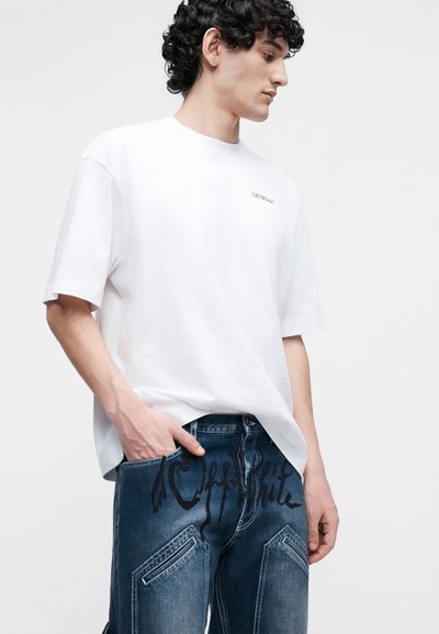 Jeune homme aux cheveux bouclés portant un T-shirt blanc surdimensionné et un jean bleu foncé avec un texte graphique noir sur la zone de la poche avant.