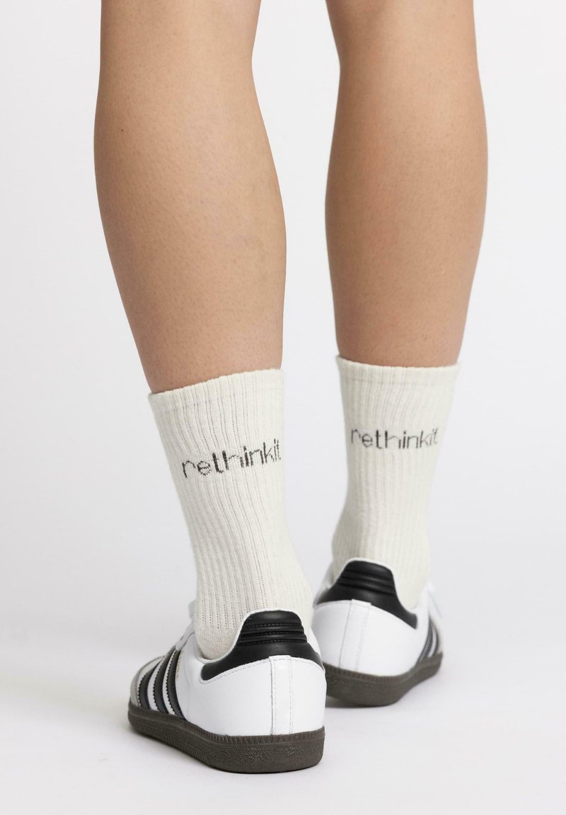 Baskets blanches avec des accents noirs et des semelles en caoutchouc texturées, associées à des chaussettes côtelées blanc cassé portant le texte « rethinkit. »