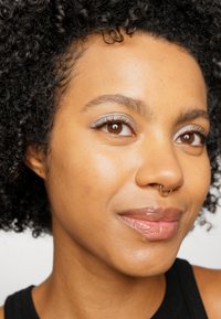 Close-up van een gezicht met mediumbruine huid, krullend zwart haar, bruine ogen en een gouden septumring. De huid heeft een gladde, glanzende textuur.