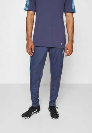 Homme portant un pantalon de sport Adidas bleu marine et un t-shirt bleu marine assorti, debout avec des crampons noirs sur un sol gris clair.