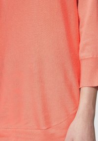 Korallfarbener Strickpullover mit gerippter Textur, Dreiviertelärmeln und abgerundetem Saum. Die Ärmelbündchen sind leicht umgeschlagen.