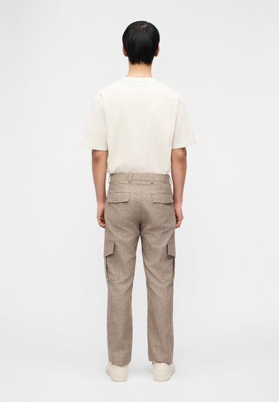 Άνδρας με κοντά μαύρα μαλλιά στέκεται με την πλάτη, φορώντας λευκό μπλουζάκι, μπεζ παντελόνι cargo με τσέπες και λευκά αθλητικά παπούτσια, μπροστά σε λευκό φόντο.