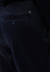 Κοντινό πλάνο σε σκούρο μπλε παντελόνι που δείχνει την πίσω τσέπη με κουμπί κα ι μικρή μεταλλική λεπτομέρεια λογότυπου κοντά στη μέση.