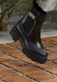 Svarte lær ankelstøvler med en kraftig gummisåle, elastiske sidestykker og et gullfarget detalj med "GUESS"-merke.