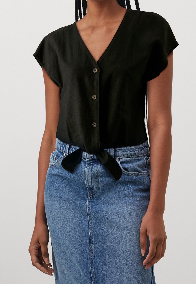 Femme portant un haut noir à manches courtes boutonné, noué à la taille, et une jupe en jean bleue taille haute.