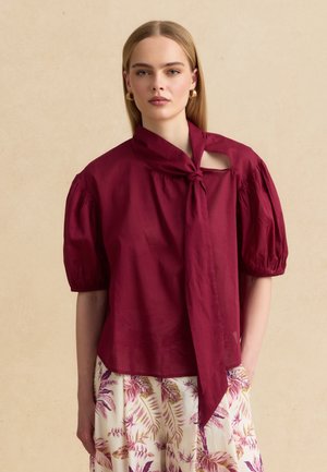 Woman with straight blonde hair wearing a burgundy puff-sleeve blouse with a necktie and floral print skirt against beige background.