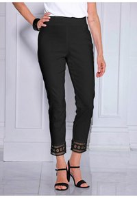 Zwarte broek met een getailleerde taille en enkel lengte, met uitgesneden kanten accenten aan de zoom. Gemaakt met zwarte hakken met bandjes.