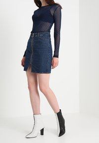 Top in rete a pois navy con maniche lunghe abbinato a una gonna di jeans scura con zip frontale e spacco, completato da stivaletti neri e bianchi.