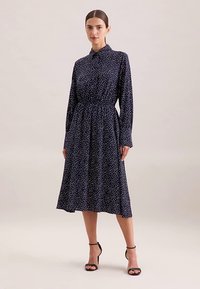 Granatowa sukienka midi w białe kropki, z kołnierzykiem zapinanym na guziki, długimi rękawami, dopasowaną talią i rozkloszowanym dołem. Noszona z czarnymi sandałami na paski.