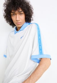 Camisa polo Nike em branco com detalhes em azul claro. Apresenta colarinho, mangas curtas e um logo no peito e na manga. Textura suave.