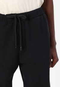 Črne trenirke iz mehkega blaga, z elastičnim pasom in vrvico, stranskimi žepi ter sproščenim krojem. Vidna teksturirana površina.