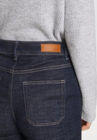 Denim jeans in dark blue with a textured finish, featuring a brown leather patch labeled "ESPRIT" on the back, and standard back pockets.