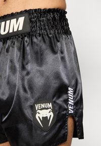 Calções pretos de cetim com cós elástico texturizado, fendas laterais e um emblema Venum com letras brancas.