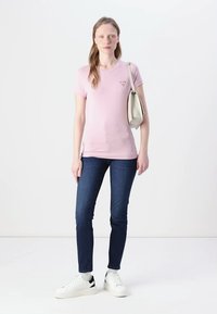 T-shirt in cotone rosa a maniche corte con un piccolo logo, abbinato a jeans in denim blu scuro e sneakers bianche con dettagli neri.