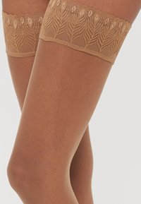 Beige augstās zeķes ar mežģīņu joslu, kas rotāta ar lapu rakstu, ar gludu tekstūru un elastīgu augšējo daļu, kas nodrošina stabilu pielāgošanu.