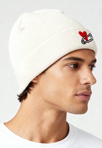 Off-white gebreide muts met geribbelde structuur, voorzien van een geborduurd rood hart en veiligheidsspeldontwerp aan de voorkant.