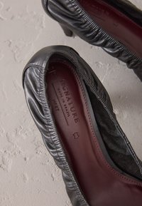 Black leather heeled shoes with gathered detailing at the ankle, smooth texture, and a burgundy insole featuring embossed branding.
